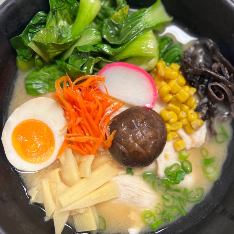 Close-up of a bowl of Ramen Mama ramen with bok choy, egg, carrots, corn, mushrooms, and scallions.